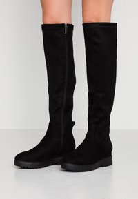 Bottes noires mi-cuisse en daim, dotées d'une fermeture éclair latérale, d'une semelle épaisse et d'une texture lisse avec un design minimaliste.