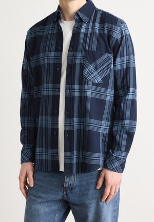Hombre que lleva una camisa de cuadros azul y marino abotonada sobre una camiseta blanca y vaqueros azul claro, de pie contra un fondo liso.