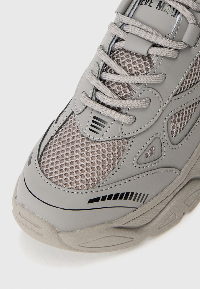 Sneaker grigio chiaro con dettagli in mesh, accenti texturizzati e strisce nere; presenta una suola testurizzata e lacci grigi per una vestibilità sicura.