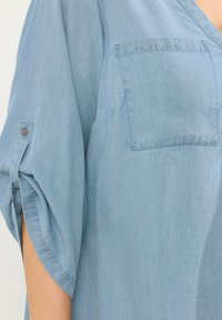 Lyseblå denimskjorte med V-hals, korte ærmer og brystlomme. Stoffet har en blød tekstur og let glans. Knapdetaljer på ærmerne.