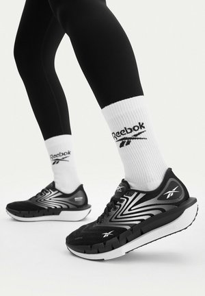 Person, die schwarze und weiße Reebok-Laufschuhe, weiße Reebok-Kurzsocken und schwarze Leggings trägt, steht vor einem weißen Hintergrund.