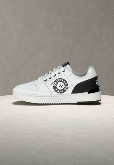 Бяло и черно ниско спортно обувки Versace Jeans Couture с лого на страничната част и петата, с връзки, поставени на светла повърхност на сив фон.