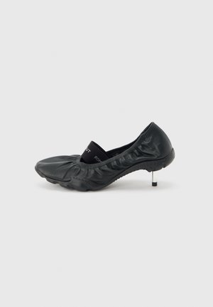 Black leather ballet flat with gathered details, elastic strap across the top, and a slim metallic kitten heel on a white background.
