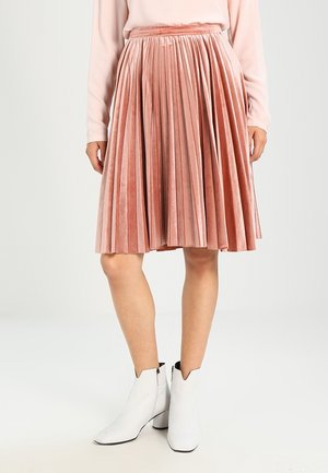 Femme portant une jupe plissée en velours rose jusqu'aux genoux, un haut à manches longues rose clair et des bottines blanches à talons carrés.