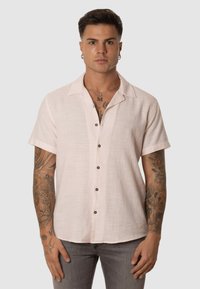 Camisa de manga corta, con cuello, en lino rosa claro con botones marrones y una textura sutil, que presenta un detalle de logo en el lado izquierdo del pecho.