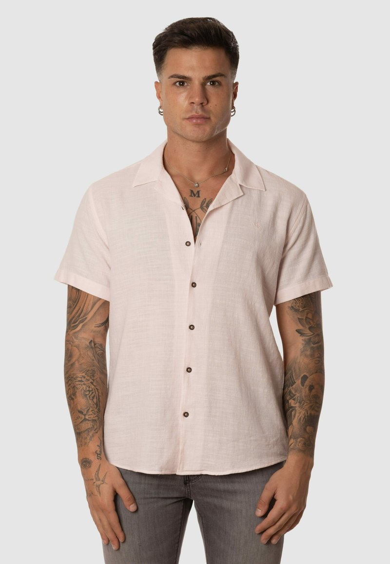 Camisa de manga corta, con cuello, en lino rosa claro con botones marrones y una textura sutil, que presenta un detalle de logo en el lado izquierdo del pecho.