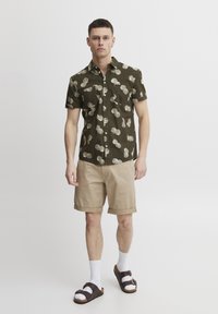Camisa de botón en verde oscuro con patrón de conchas blancas, combinada con pantalones cortos beige, calcetines blancos y sandalias marrones. Mangas cortas, corte relajado.