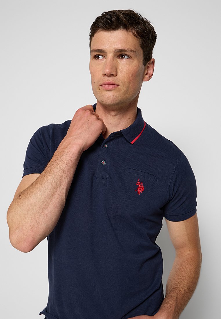 Navyblaues Poloshirt aus atmungsaktivem Material, mit rotem Kragen und kleinem rotem Logo auf der Brust, mit einer strukturierten Oberfläche.
