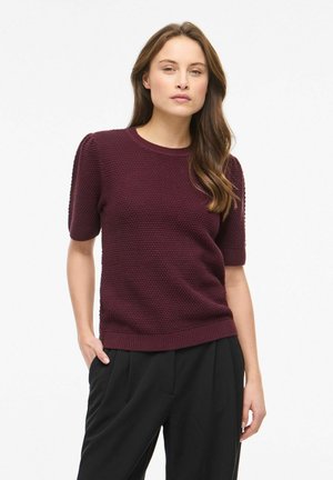 Femme portant un pull bordeaux à manches courtes texturé et un pantalon noir plissé, debout avec une main dans la poche, sur fond blanc.