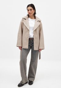 Beež villasegu jakk laia krae, kahe nööbi ja külgedel sidemete ning hallide teksade ja mustade kingadega. All on lihtne valge T-särk.