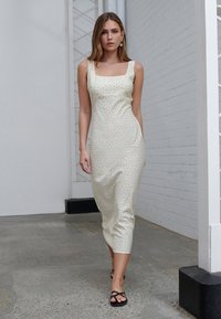 Knie-langes, cremifarbenes Kleid mit quadratischem Ausschnitt und verstreuten schwarzen Punkten, aus glattem Stoff; kombiniert mit schwarzen Riemchensandalen.