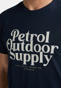 Námořnická modrá bavlněná tričko s velkým, tučným bílým textem "Petrol Outdoor Supply" a s dalším menším zeleným textem pod ním.