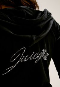 Hoodie preto em velour com um texto decorativo "Juisy" em strass prateados na parte de trás, com um capuz suave e texturizado.