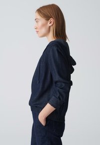 Giacca di jeans con cappuccio di colore blu scuro, caratterizzata da una vestibilità comoda, polsini elasticizzati e una tasca a marsupio. Texture morbida con dettagli di cucitura minimi.