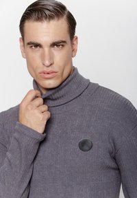 Szary sweter z dekoltem typu golf, z prążkowanej tkaniny, z czarną skórzaną łatą logo po lewej stronie, wykonany z miękkiego, teksturowanego materiału z wyraźnym wykończeniem przy szyi.