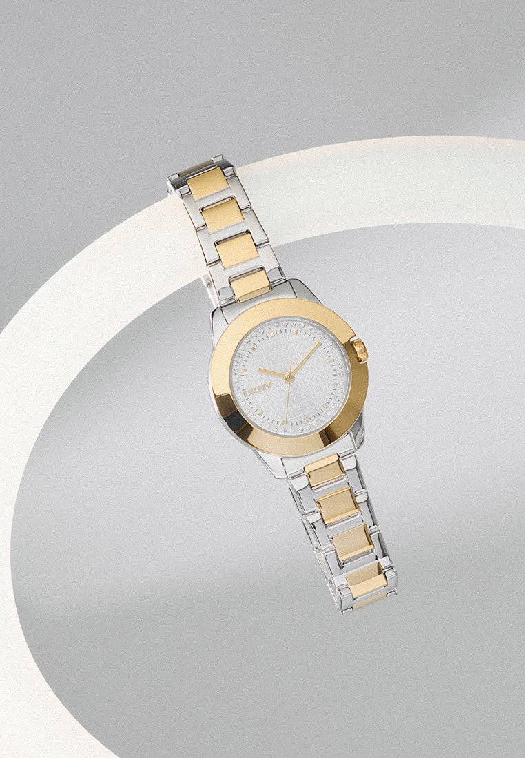 Montre-bracelet bicolore en argent et or avec un cadran blanc à motif et une lunette dorée, présentée sur une surface blanche incurvée avec un arrière-plan gris.