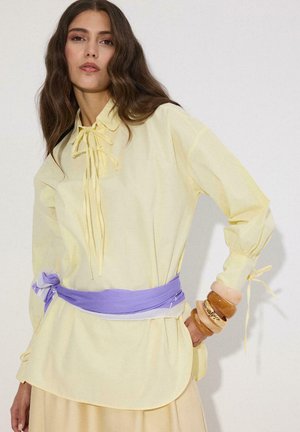 Γυναίκα που φοράει μπλούζα σε φωτεινό κίτρινο χρώμα με δεμένο λαιμόκοψη, ζώνη λεβάντας και στοίβα από καφέ και μπεζ βραχιόλια στον καρπό της.