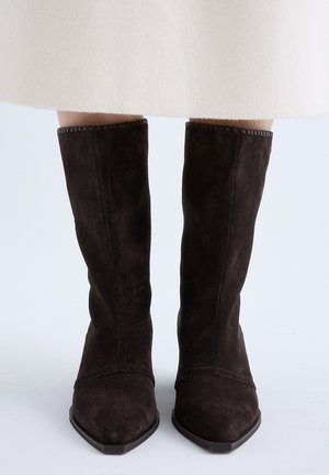 Par de botas de ante marrón oscuro hasta media pantorrilla con puntera puntiaguda, usadas con una falda de color claro sobre un fondo blanco.