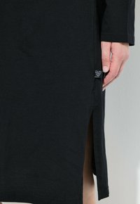 Schwarzes Baumwollkleid mit langen Ärmeln und seitlichen Schlitzen; besitzt ein kleines Etikett auf der linken Seite. Glatte Textur, schlichtes Design.