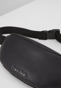 Czarna skórzana nerka o gładkiej fakturze, z wyraźnym srebrnym logo "Calvin Klein" oraz czarnym regulowanym paskiem.