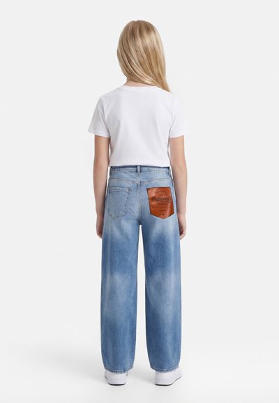 Child with long blonde hair stands facing away, wearing a white t-shirt, wide-leg faded blue jeans with a brown leather patch, and white sneakers.