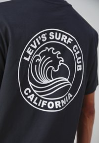 T-shirt noir avec un graphisme circulaire blanc représentant des vagues stylisées et le texte "Levi's Surf Club California." Matière en coton, col rond.