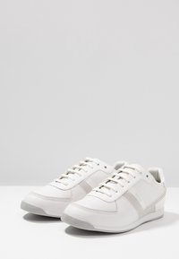 Bílé kožené tenisky s texturovanými šedými akcenty, nízkým profilem, kulatou špičkou a plochou gumovou podrážkou. Obsahují šněrování a detaily loga.