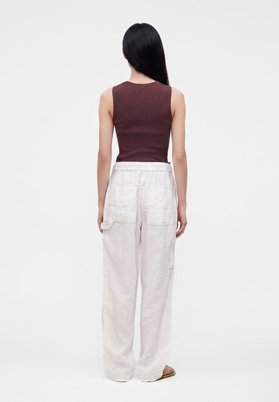 Personne aux longs cheveux noirs portant un haut marron sans manches et un pantalon blanc ample avec de grandes poches à l'arrière, debout de dos.
