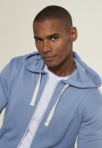 Hellblaues Zip-Hoodie mit einer Kordelzugkapuze, beigen Akzenten an den Kordeln und einem weißen T-Shirt darunter. Weiche Textur.