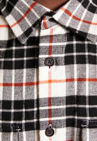 Camisa de franela a cuadros en blanco y negro con acentos rojos. Cuenta con un cierre de botones, cuello y una textura suave.