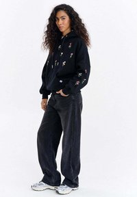 Černá mikina s kapucí s květinovou výšivkou v různých barvách, kombinovaná s volnými černými džínami a bílými sportovními botami. Hladká textura látky.