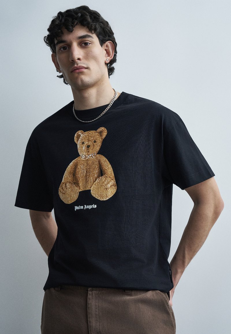 Junger Mann mit lockigem Haar, der ein schwarzes T-Shirt mit einem braunen Teddybär-Motiv und einer silbernen Kettenhalskette trägt, vor einem einfarbigen Hintergrund.