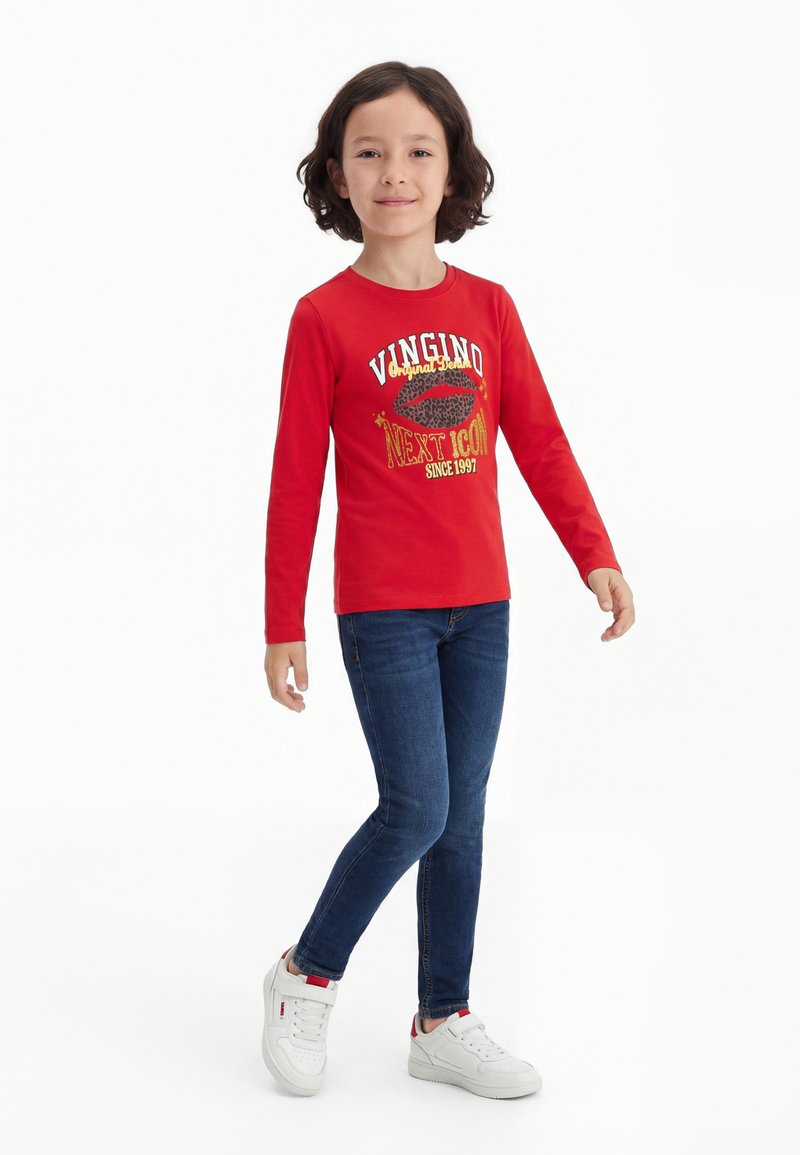 Bambino con capelli corti e scuri che indossa una maglietta rossa a maniche lunghe con scritta, jeans blu scuro e scarpe da ginnastica bianche, in piedi su sfondo bianco.