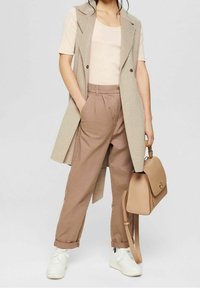 Beige bezrukávový kabát přes světlo barevný top, v kombinaci s volnými hnědými kalhotami a bílými teniskami. Drží světle hnědou kabelku se strukturovaným designem.