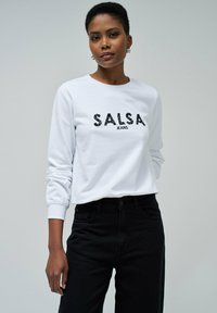 Sudadera blanca con "SALSA JEANS" estampado en lentejuelas negras, combinada con jeans negros. La tela parece suave con puños ajustados.