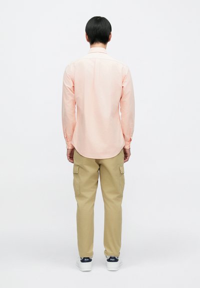 Ροζ ανοιχτόχρωμο πουκάμισο με μακριά μανίκια και καμπύλη γραμμή στη βάση, συνδυασμένο με μπεζ cargo παντελόνια με πλαϊνές τσέπες και λευκά αθλητικά παπούτσια με ναυτικό μπλε λεπτομέρειες.