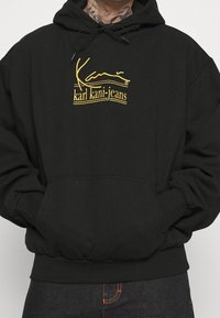 Svart huvtröja i mjukt tyg, med en gul logotyp och text på framsidan, med en känguruficka och ribbade ärmslut.