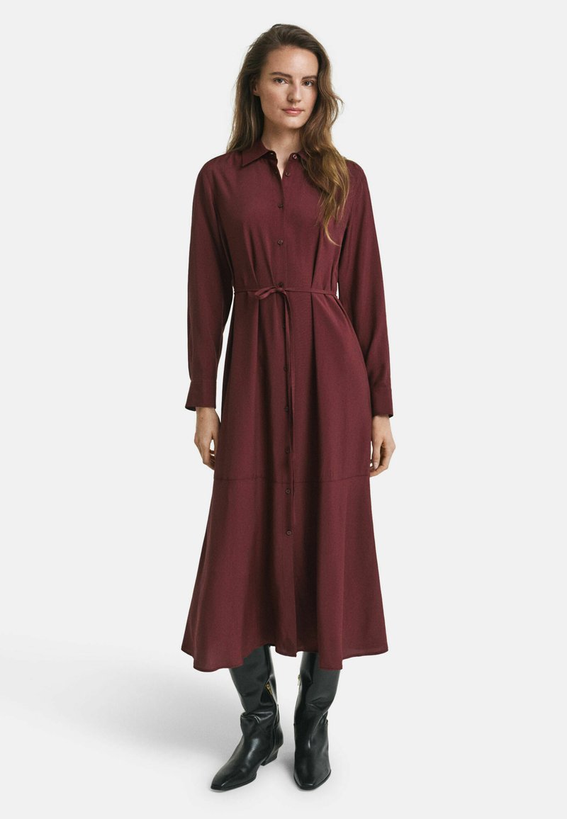 Burgunderfarbenes Langarmkleid mit Knopfleiste und Bindegürtel, das eine fließende Silhouette und eine Länge bis zur Mitte der Waden hat. Getragen mit schwarzen Stiefeln.