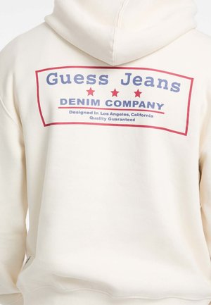 Kremna kapuca s velikim grafičkim uzorkom na leđima na kojem piše "Guess Jeans" u plavoj boji i "DENIM COMPANY" u crvenoj boji, ukrašena sa tri crvene zvijezde.