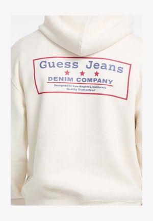 Kremna kapuca s velikim grafičkim uzorkom na leđima na kojem piše "Guess Jeans" u plavoj boji i "DENIM COMPANY" u crvenoj boji, ukrašena sa tri crvene zvijezde.
