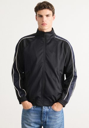 Svart zip-up sportsjakke med hvit pipingen og elastiske mansjetter, kombinert med lyseblå jeans. Glatt stoff, slank design.