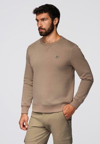 Beige mikina s dlouhými rukávy, kulatým výstřihem, žebrovanými manžetami a lemem. Obsahuje malou značkovou nášivku na hrudi. Hladká textura.