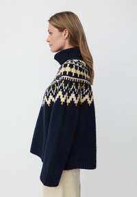 Granatowy sweter wełniany z wysokim prążkowanym kołnierzem oraz geometrycznymi wzorami w kolorze kremowym i żółtym na górnej części. Luźny krój, miękka tekstura.