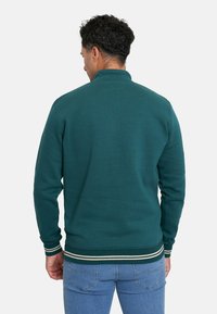 Suéter verde oscuro con un cuello alto, puños acanalados y detalle de ribete a rayas. Textura suave en un corte relajado.
