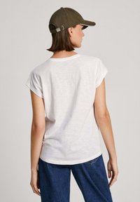 Camiseta blanca de manga corta con cuello redondo y ajuste relajado, combinada con jeans de mezclilla azules. Una gorra verde completa el look.