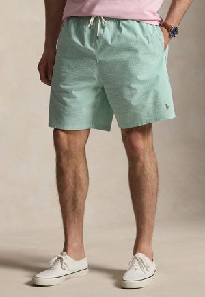 Bărbat purtând pantaloni scurți verzi și albi cu dungi, cu șnur, tricou roz, ceas și adidași albi cu șireturi, stând cu mâinile în buzunare.