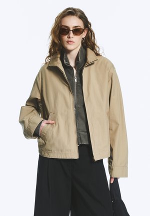 Woman wearing beige jacket over gray zip-up sweater, black wide pants, brown sunglasses, holding a black bag against a white background.