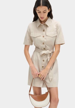 Kobieta ubrana w beżową, krótko rękawową sukienkę z paskiem i przednimi kieszeniami, trzymająca przed sobą małą brązowo-kremową torebkę.