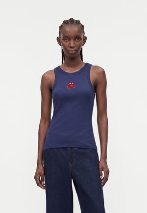 Femme aux cheveux tressés porte un débardeur bleu marine avec un petit motif crabe rouge, associé à un jean large foncé, debout devant un fond uni.