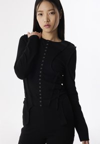 Schwarzes Langarmoberteil mit strukturiertem Design, ausgestattet mit silbernen Druckknöpfen an der Vorderseite und asymmetrischen Seitenakzenten. Glatte Stoffoberfläche.